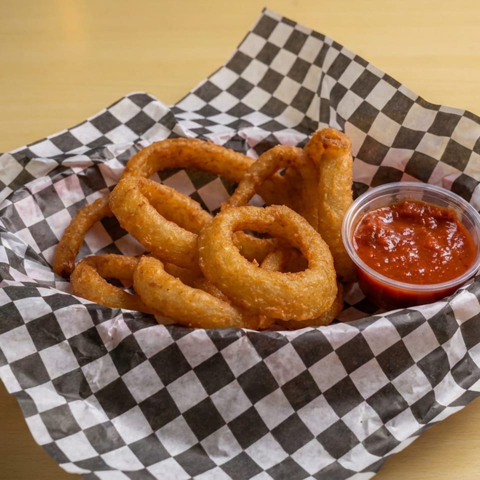 Crispy golden onion rings served in checkered paper with a side of red dipping sauce on a wooden table.