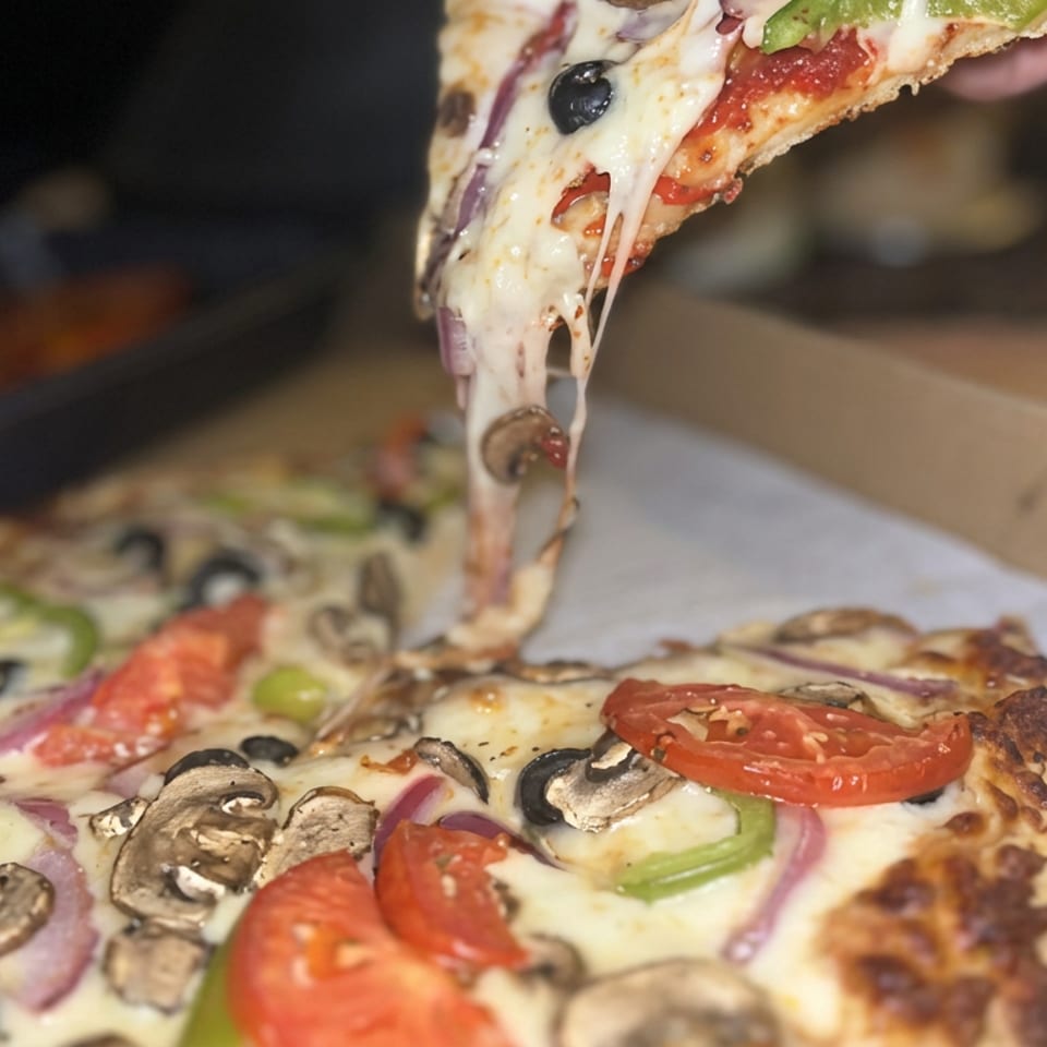Hand lifts a cheesy vegetable pizza slice from an open cardboard box, revealing mushrooms, tomatoes, olives, peppers, and onions.