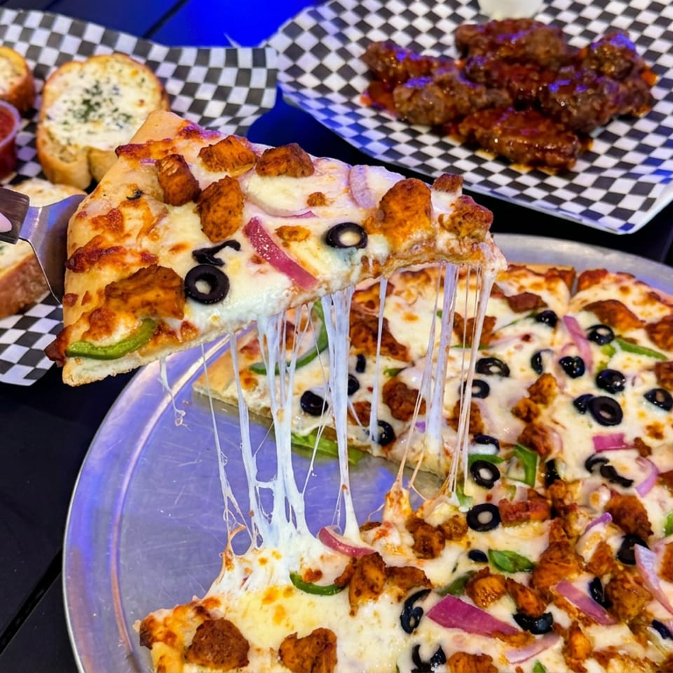 A hand lifts a cheesy pizza slice topped with olives, peppers, onions, and chicken, with garlic bread and saucy wings behind.